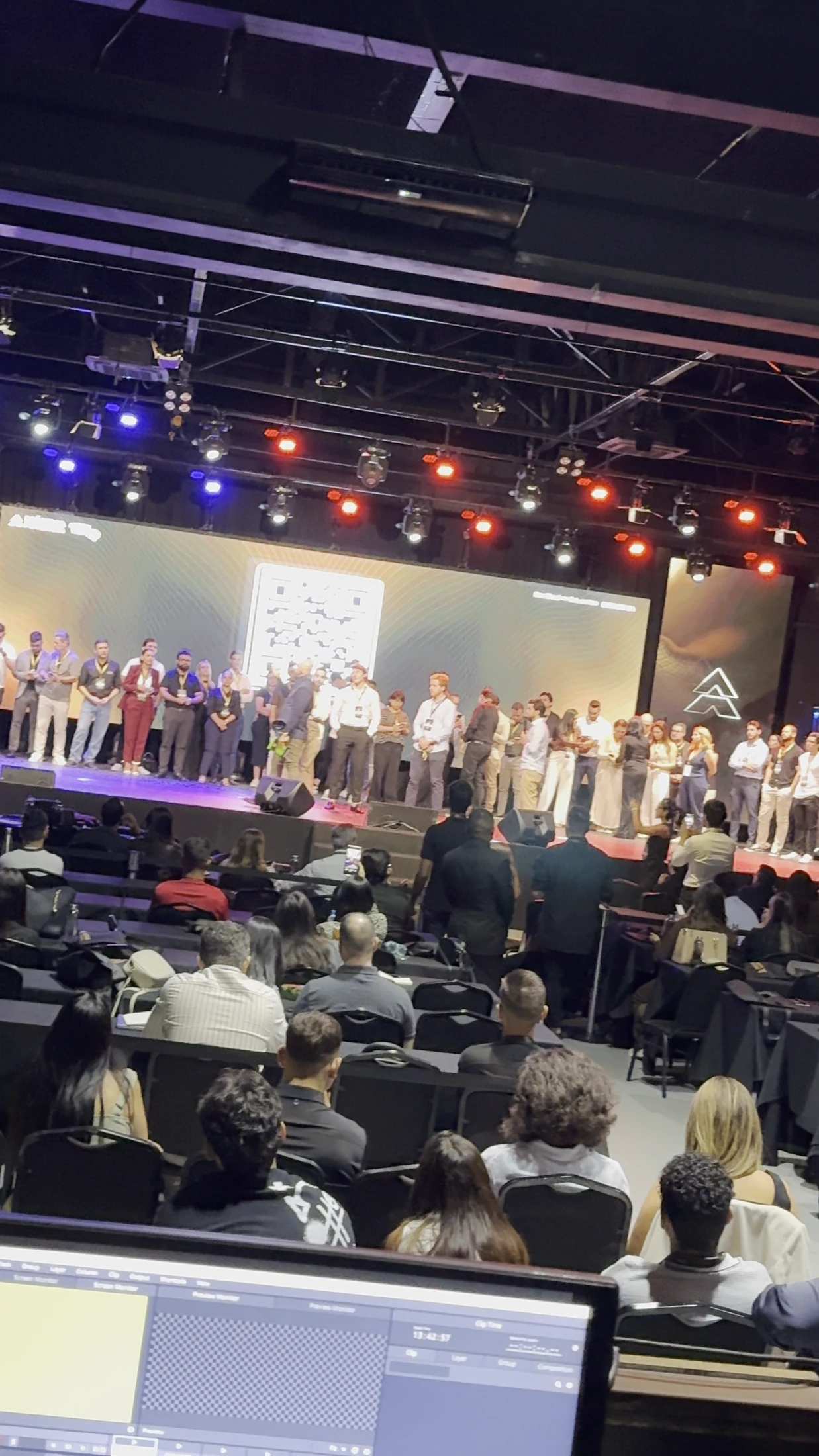 Palco com telões, iluminação cênica e público em convenção
